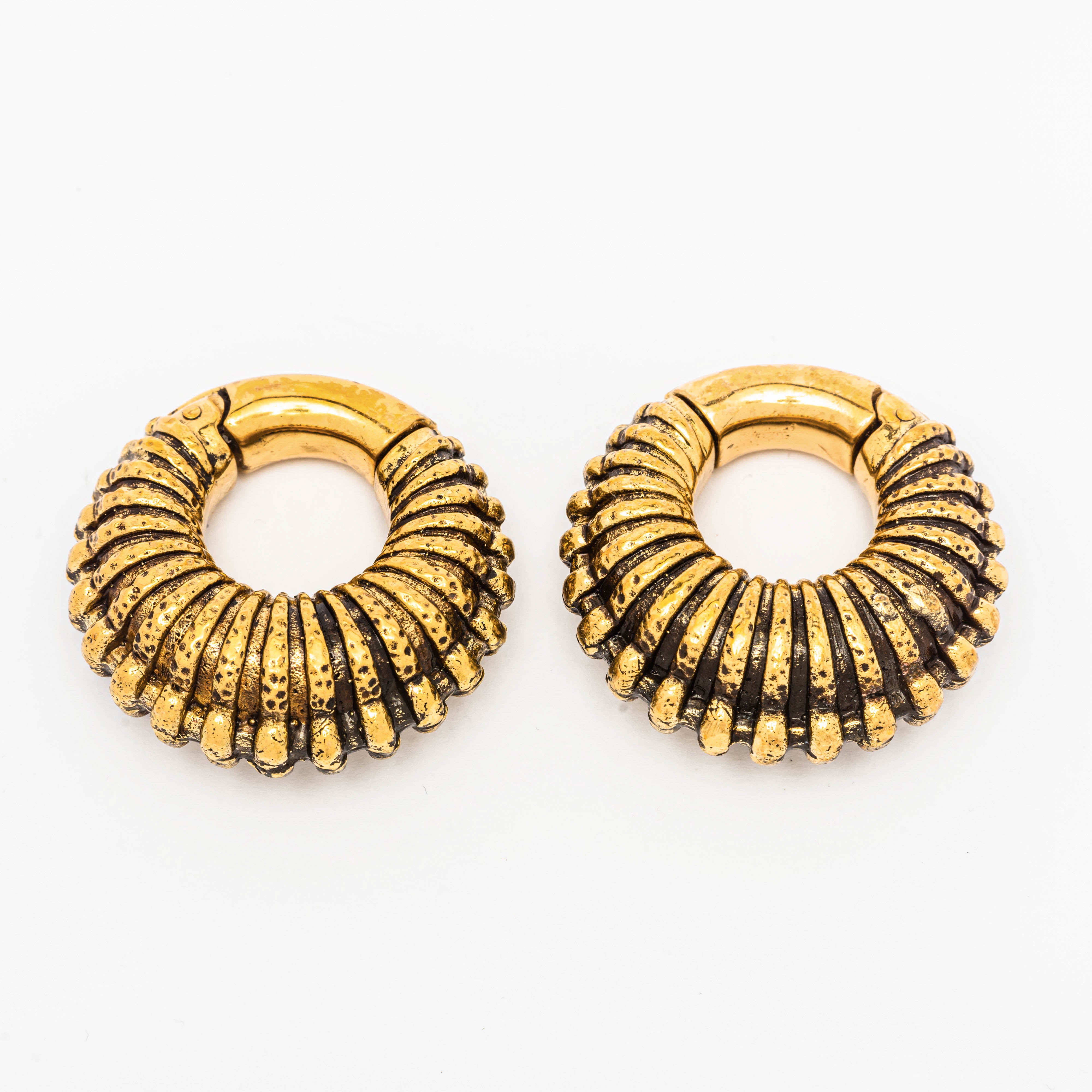 Introducing the TRILOBITE Large Biomechanical Clicker Ear Weights in Gold Brass by Kaya Movement. These 0 gauge ear weights boast a distinctive ribbed texture and slightly oxidized finish, evoking a vintage tribal aesthetic. Their circular design with a hinge mechanism at the top ensures easy fastening. The product is showcased against a minimalist white background.